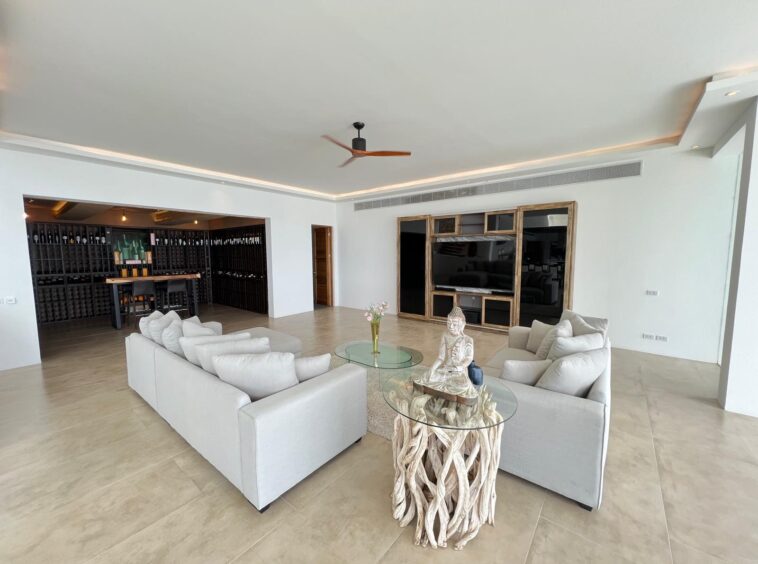 Spacious living room with white sofas, glass coffee table, and a Buddha statue centerpiece; wine cellar visible to the left and a large TV wall behind.
