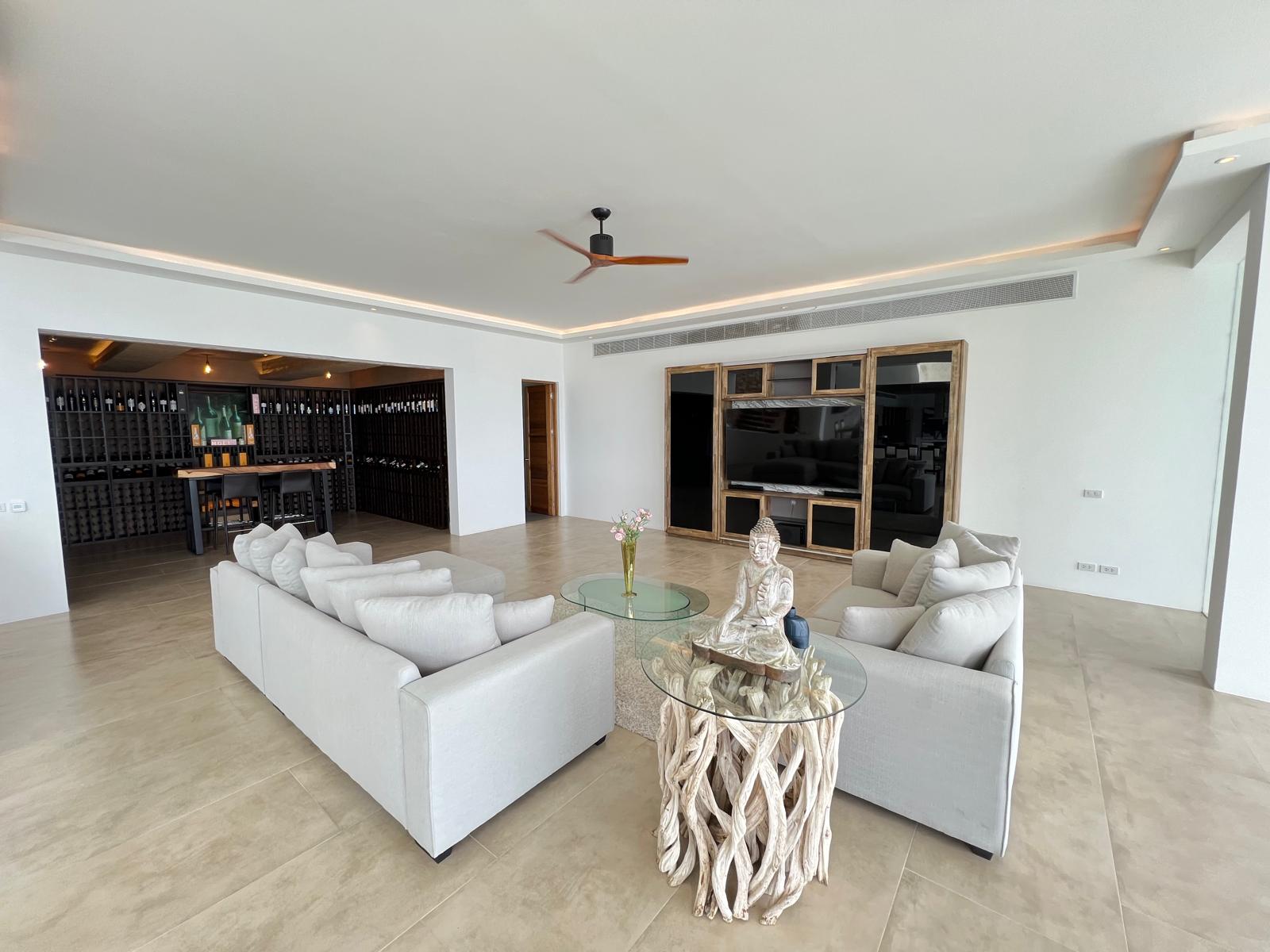 Spacious living room with white sofas, glass coffee table, and a Buddha statue centerpiece; wine cellar visible to the left and a large TV wall behind.