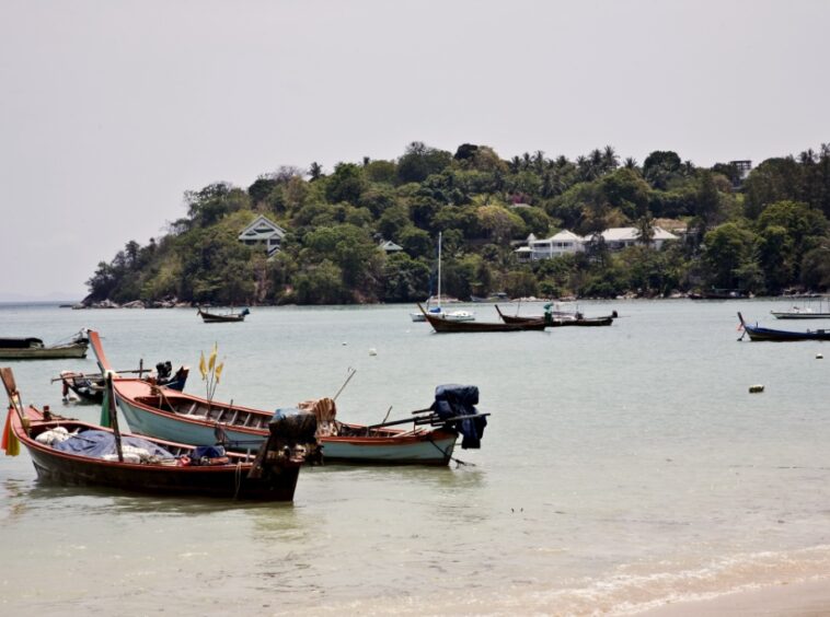 Small colorful fishing boats anchored near a light turquoise shore with a forested island and houses in the background
