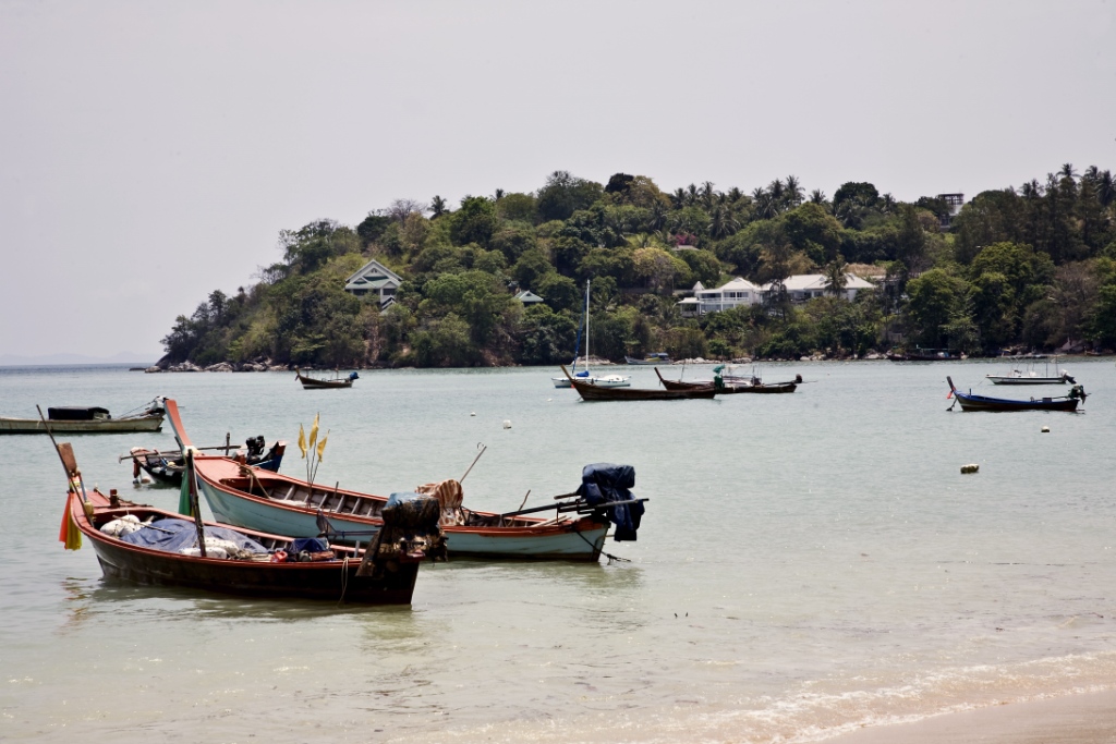 Small colorful fishing boats anchored near a light turquoise shore with a forested island and houses in the background
