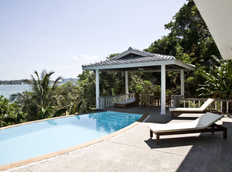 Poolside terrace with two white lounge chairs, a shaded gazebo, and tropical foliage overlooking the ocean.
