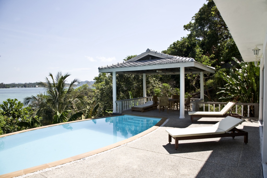 Poolside terrace with two white lounge chairs, a shaded gazebo, and tropical foliage overlooking the ocean.