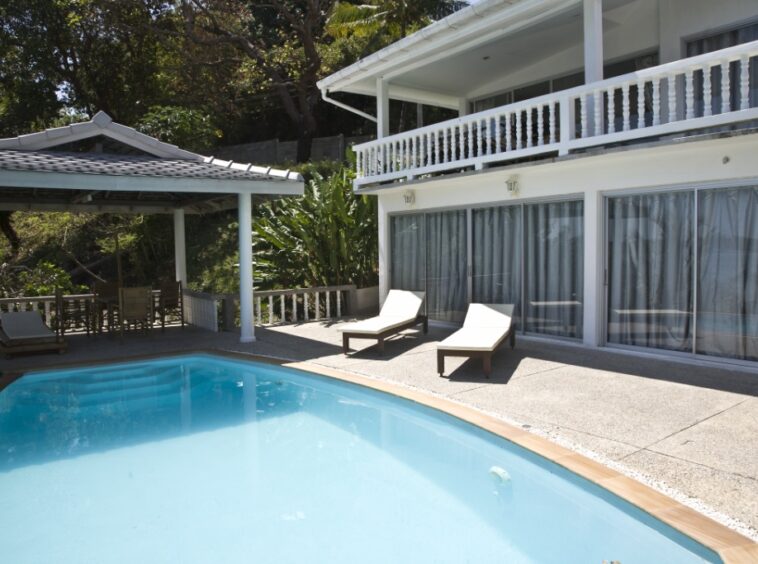 Outdoor pool area with two white lounge chairs beside glass doors of a white house and a shaded seating area under a pavilion.