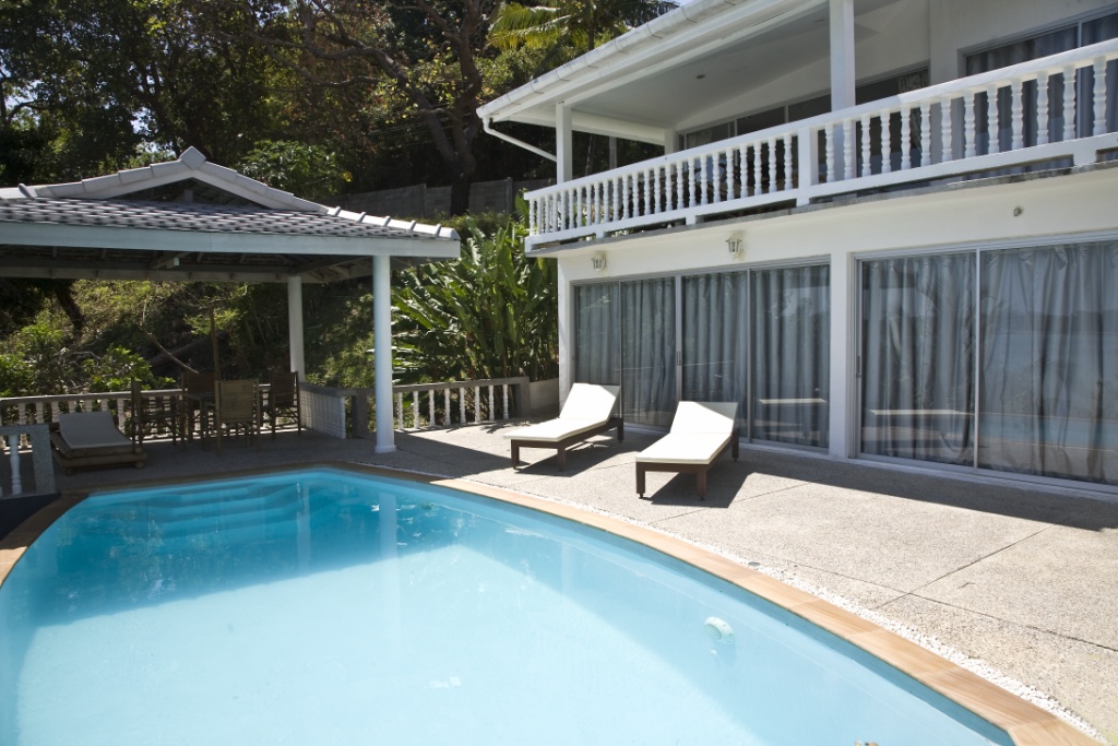 Outdoor pool area with two white lounge chairs beside glass doors of a white house and a shaded seating area under a pavilion.