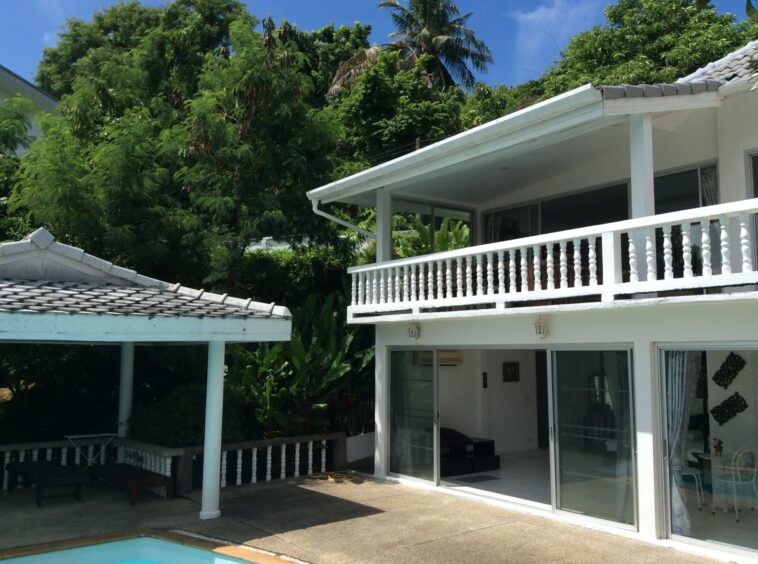 White two-story house with a glass sliding door opening to a balcony, surrounded by tropical trees and a small pool edge in the foreground.