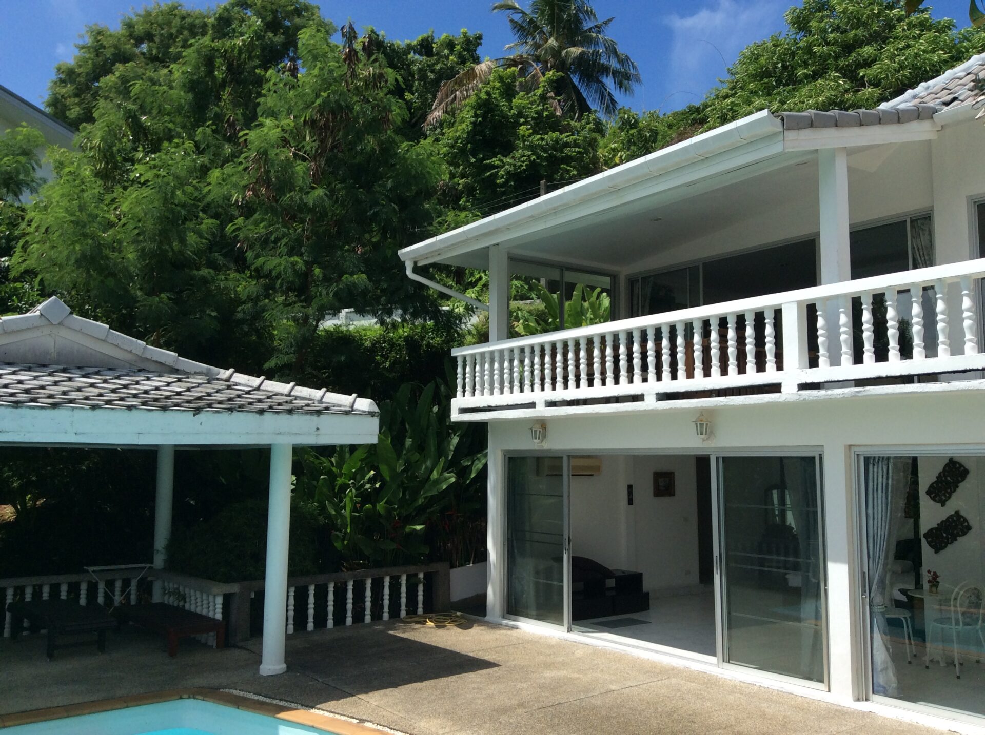 White two-story house with a glass sliding door opening to a balcony, surrounded by tropical trees and a small pool edge in the foreground.