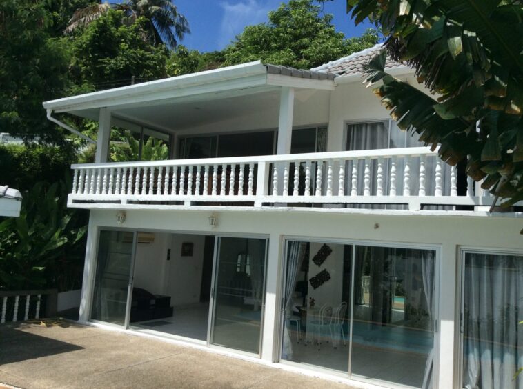 White two-story house with large glass sliding doors opening to a covered balcony, surrounded by tropical plants.