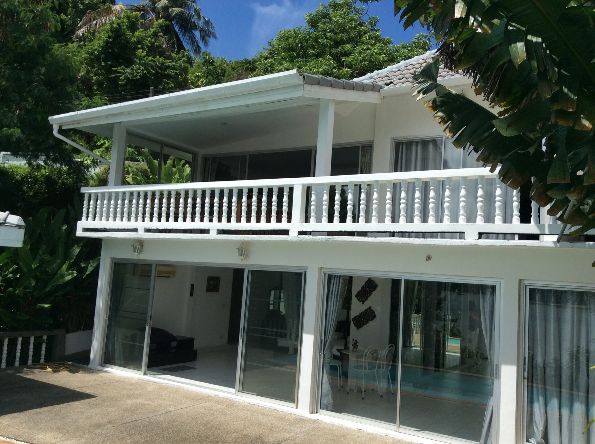 White two-story house with large glass sliding doors opening to a covered balcony, surrounded by tropical plants.