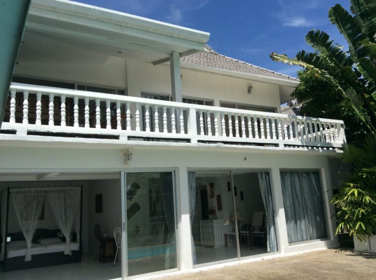 White two-story house with a covered balcony and white balustrade, sliding glass doors, and tropical plants against a clear blue sky.