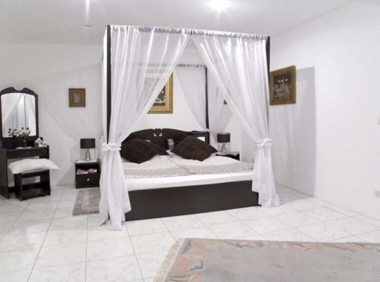 Decorative four-poster bed with sheer white canopy curtains in a bright bedroom, dark wood furniture and tiled floor.