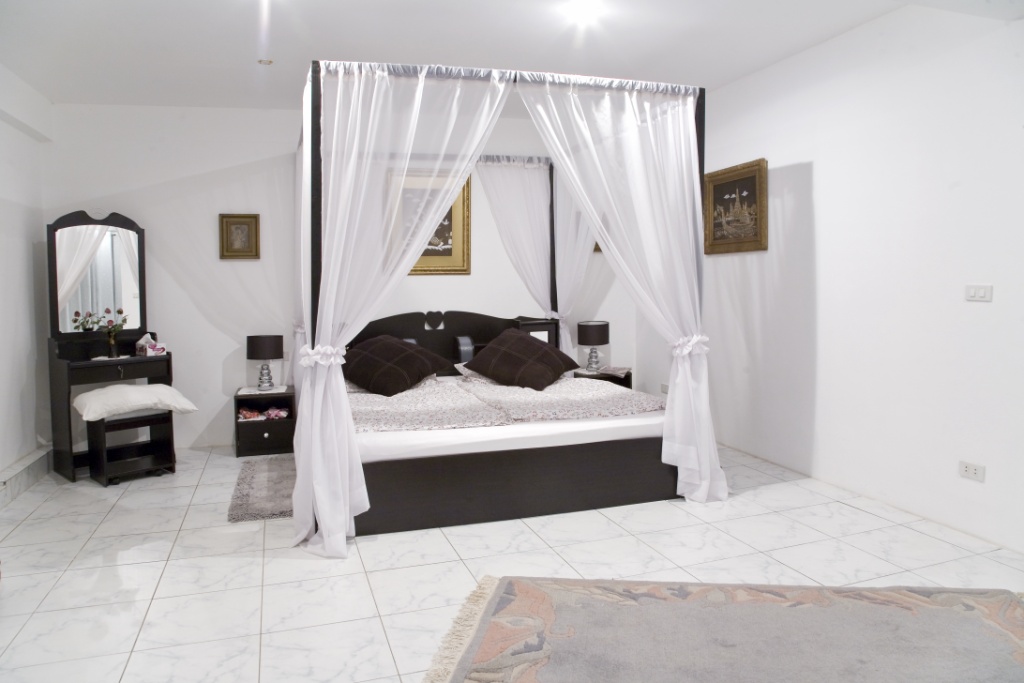 Decorative four-poster bed with sheer white canopy curtains in a bright bedroom, dark wood furniture and tiled floor.