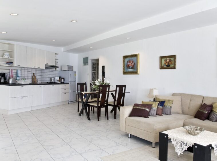 Open-plan living area with a beige sectional, glass-top dining table, and a white kitchen in the background under recessed lights.
