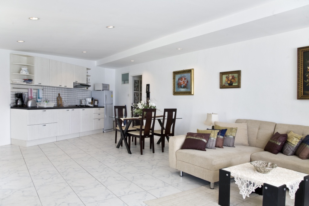 Open-plan living area with a beige sectional, glass-top dining table, and a white kitchen in the background under recessed lights.