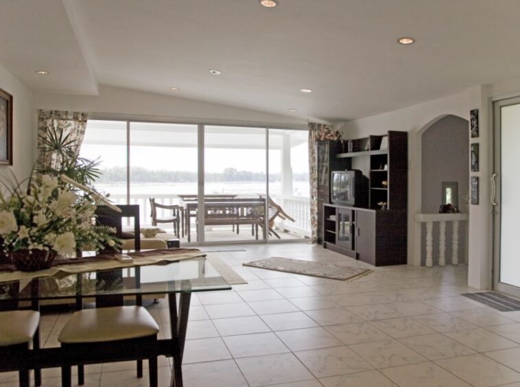 Bright open living room with a glass dining table, floral centerpiece, and a view through sliding doors to a water-facing balcony. A spacious lounge area with a TV cabinet, shelves, and tiled floor leading to a sunlit outdoor deck.