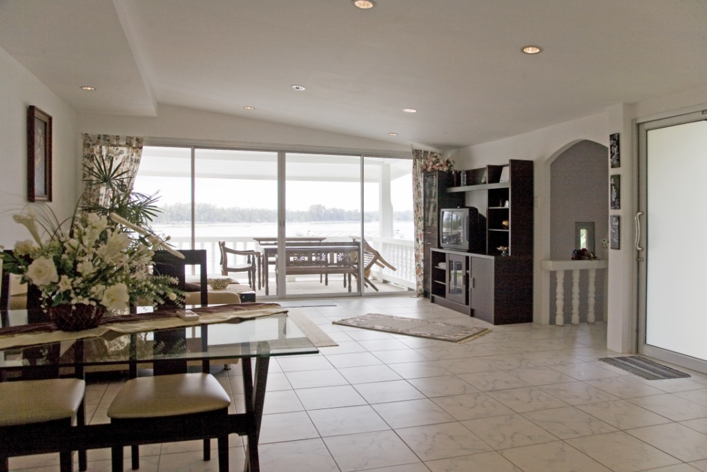 Bright open living room with a glass dining table, floral centerpiece, and a view through sliding doors to a water-facing balcony. A spacious lounge area with a TV cabinet, shelves, and tiled floor leading to a sunlit outdoor deck.