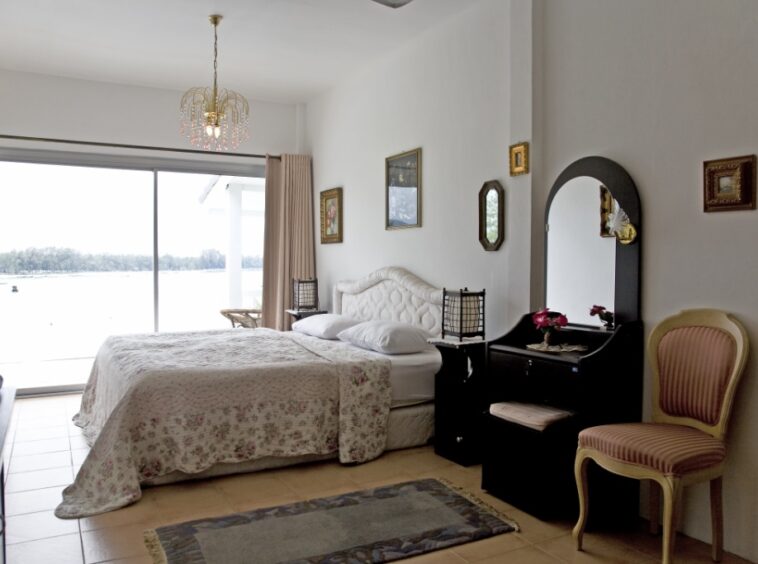 Bright bedroom with a floral quilted bed, sliding glass door to a lake view, white walls, and a black vanity with a mirror beside a pink-striped chair.