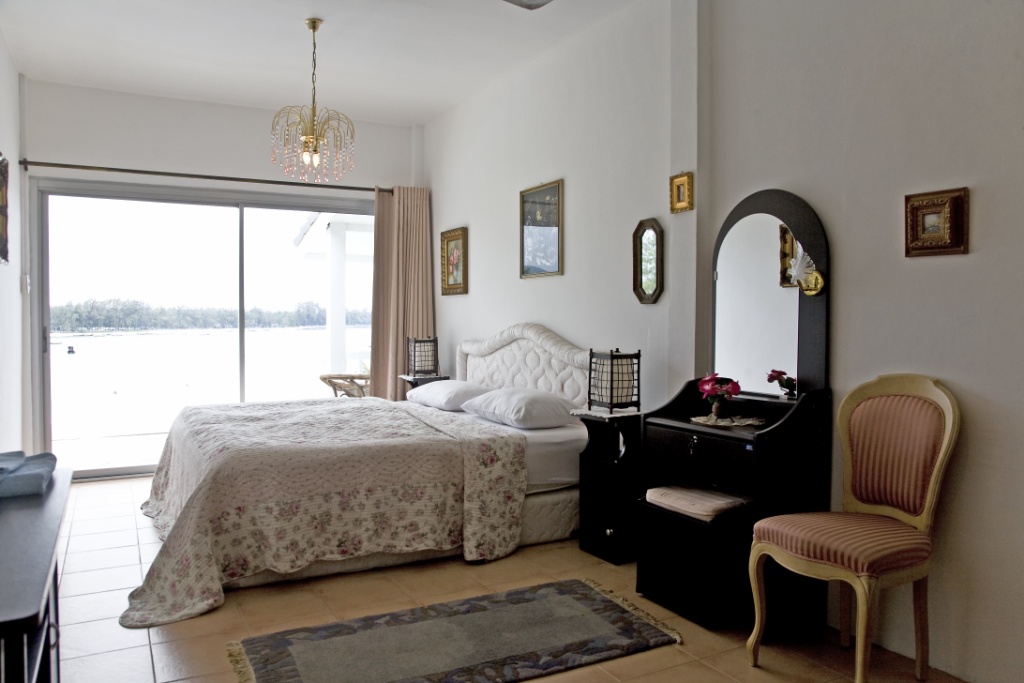Bright bedroom with a floral quilted bed, sliding glass door to a lake view, white walls, and a black vanity with a mirror beside a pink-striped chair.