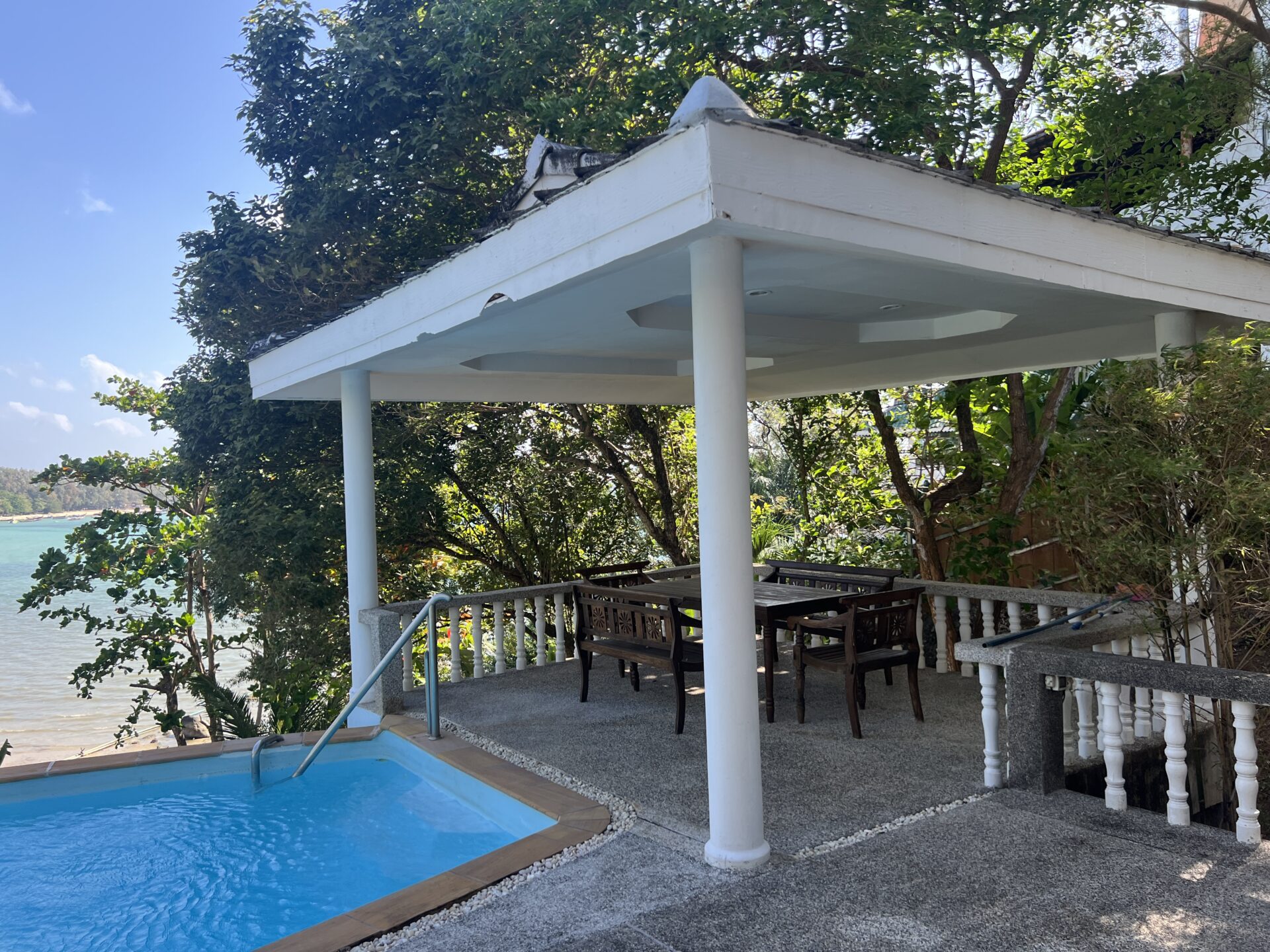Outdoor covered patio overlooking a pool and waterfront with white columns, trees, and dark wooden dining table and chairs nearby.