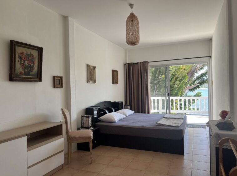 Hotel-style bedroom with two neatly made beds facing a glass door to a balcony overlooking palm trees and the ocean.
