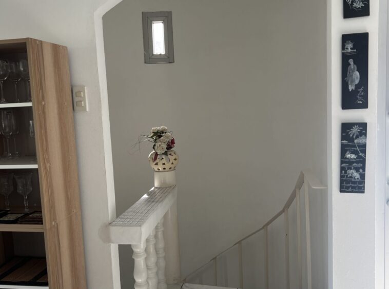 White interior stair alcove with a curved arch and a small rectangular window; a vase of flowers sits on a tiled railing, near a wooden cabinet with glassware.