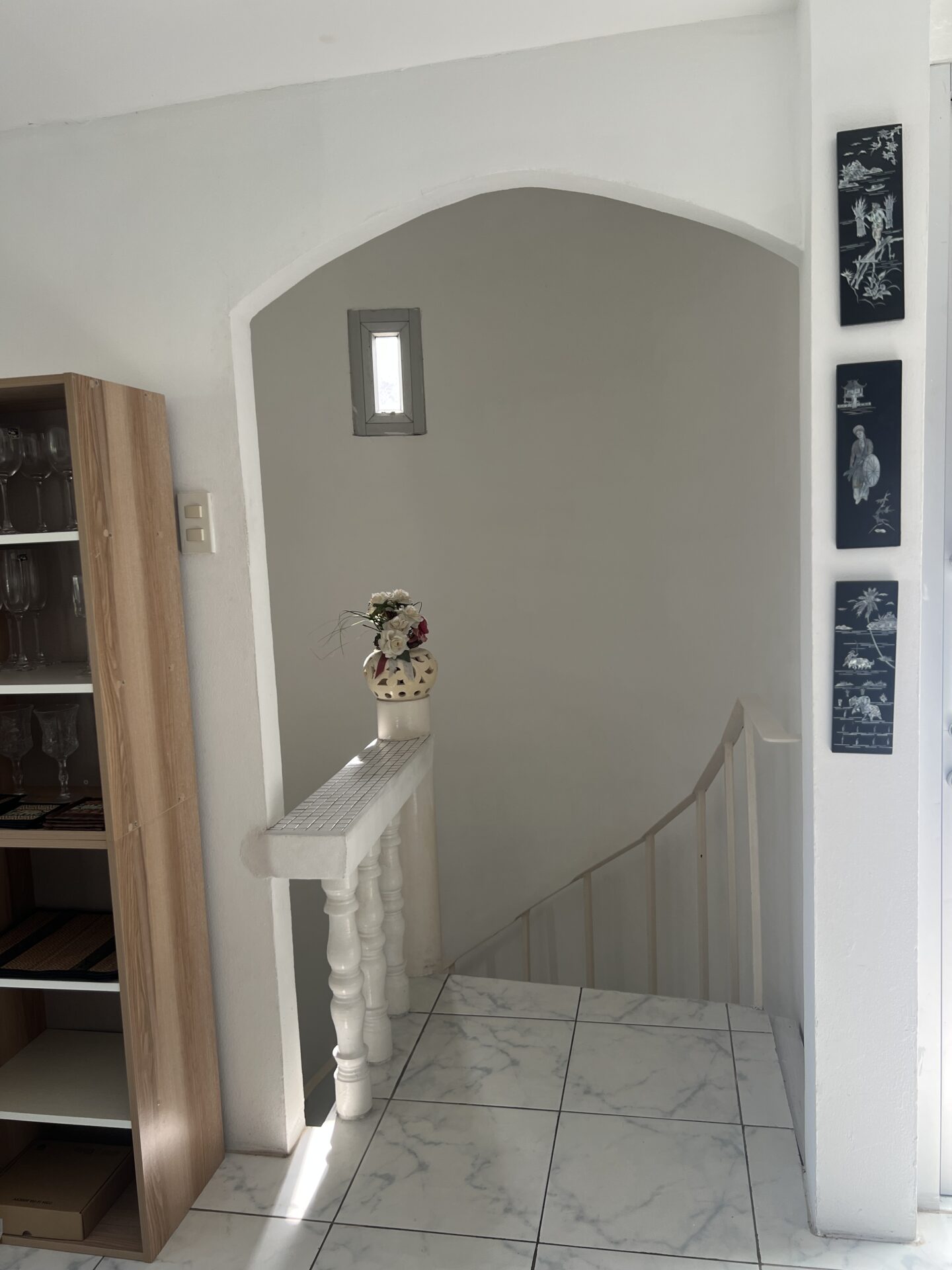 White interior stair alcove with a curved arch and a small rectangular window; a vase of flowers sits on a tiled railing, near a wooden cabinet with glassware.