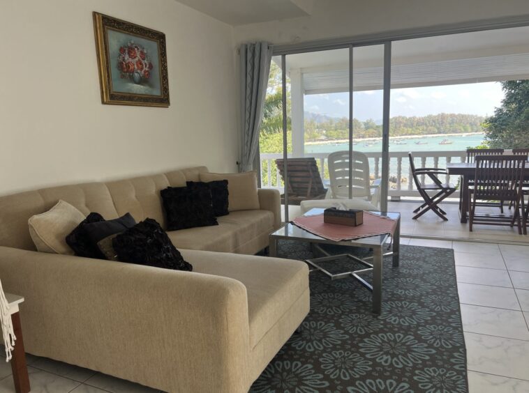 Beige sectional sofa with black throw pillows in a living room, sliding glass doors open to a balcony with a sea view and boats on the water.