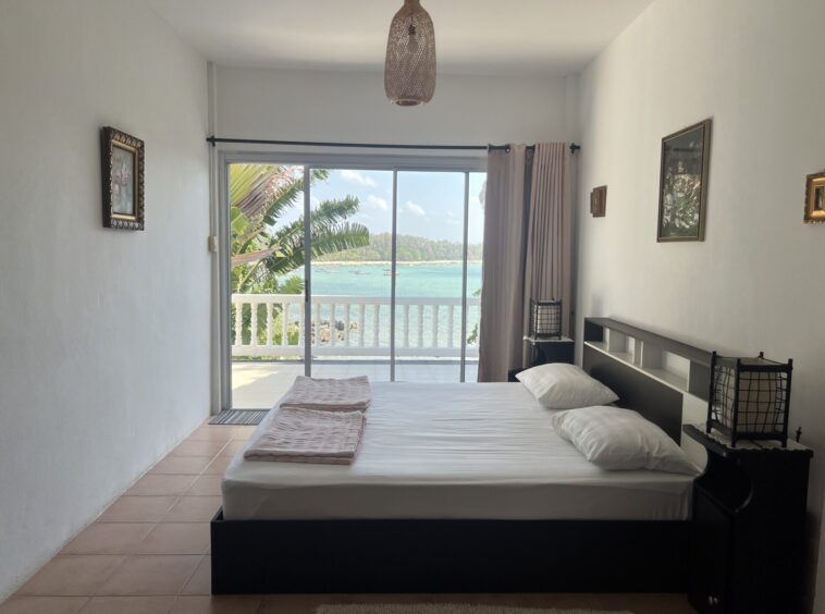 Bedroom with a large sliding glass door to a balcony overlooking the ocean; bed in foreground with pink throws and white pillows.