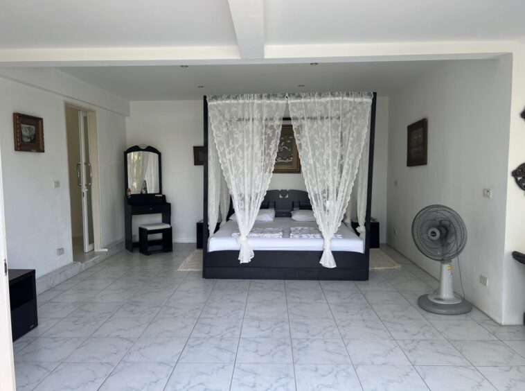 Dark four-poster bed with white lace canopy and bedding in a bright, tiled-room, with a large standing fan to the right.