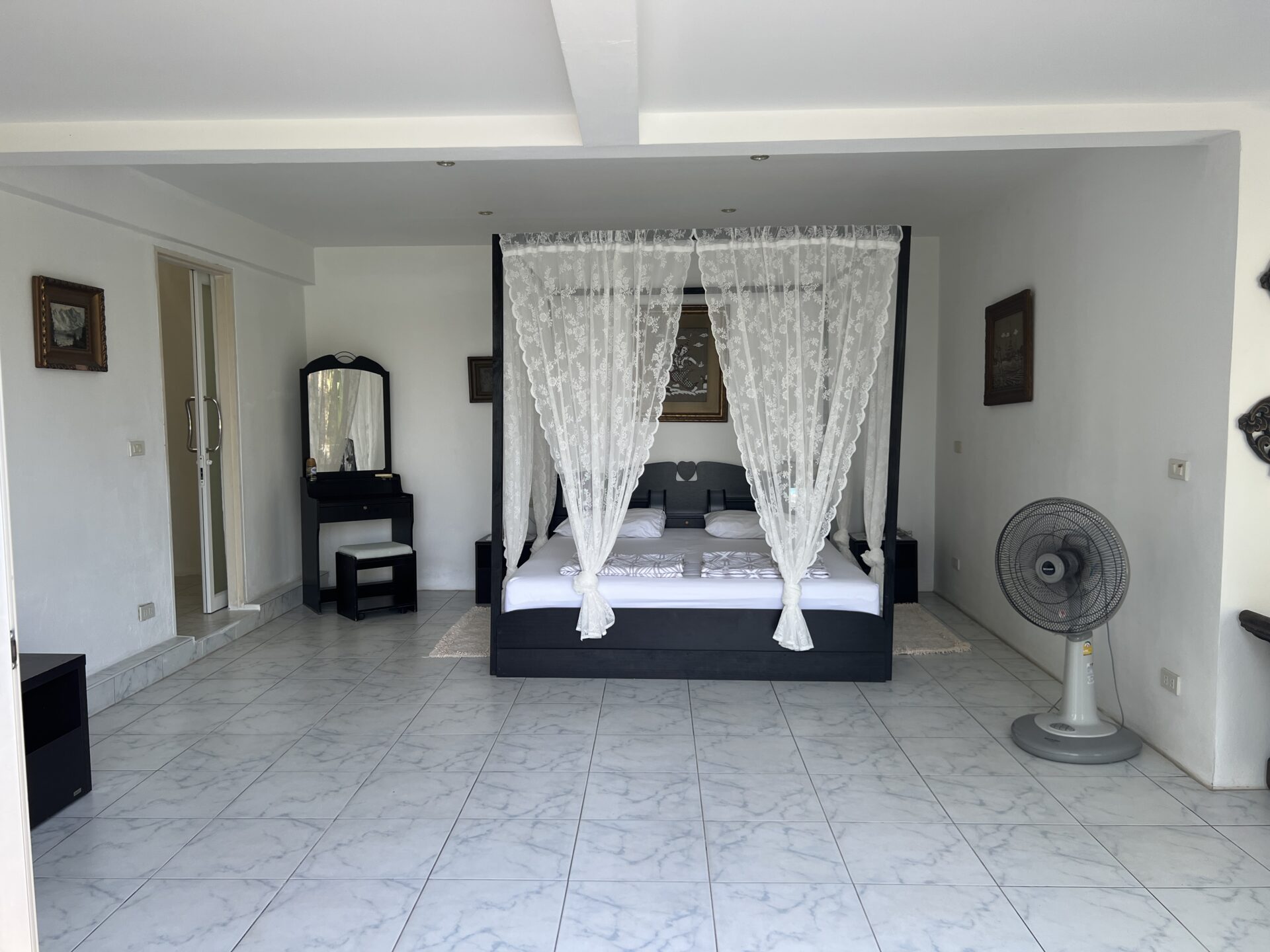 Dark four-poster bed with white lace canopy and bedding in a bright, tiled-room, with a large standing fan to the right.