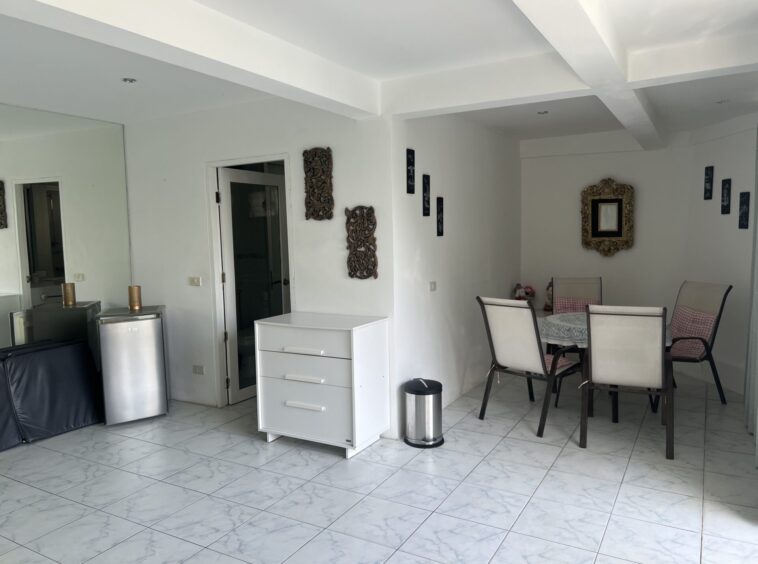 Bright dining area with a round table, six cushioned chairs, and a gold ornate mirror on white walls next to a glass cabinet and white furniture.