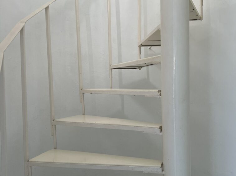 White spiral staircase with a central support column in a plain white-walled room, tiled floor visible below.