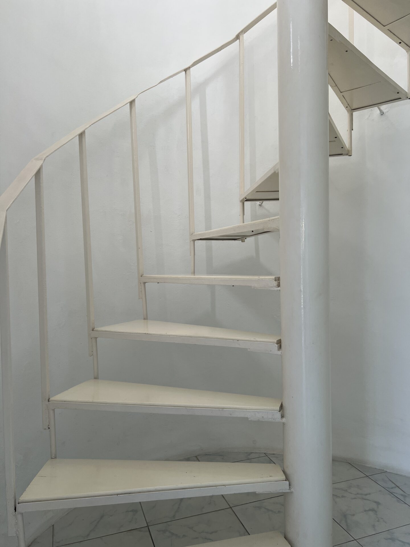 White spiral staircase with a central support column in a plain white-walled room, tiled floor visible below.