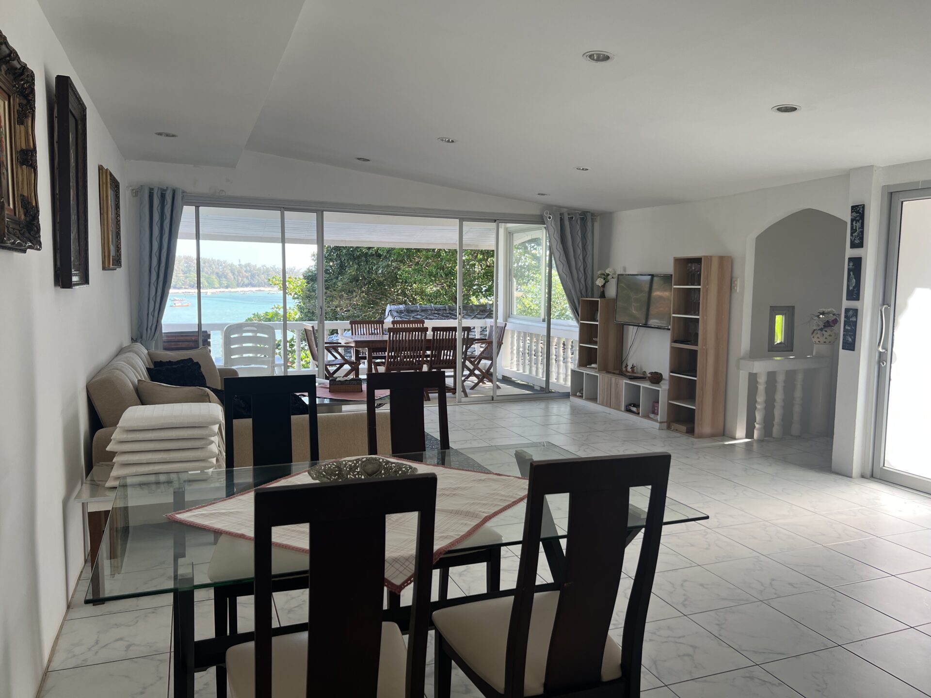Bright open-plan living and dining area with a glass dining table, dark wooden chairs, and a beige sofa, leading to a balcony with an ocean view.