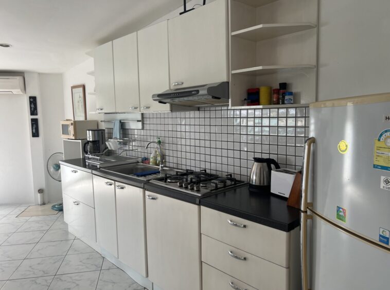 Long view of a modern kitchen with white cabinets, a black countertop, and a tiled backsplash; stove, kettle, toaster, microwave, and coffee maker on counters.