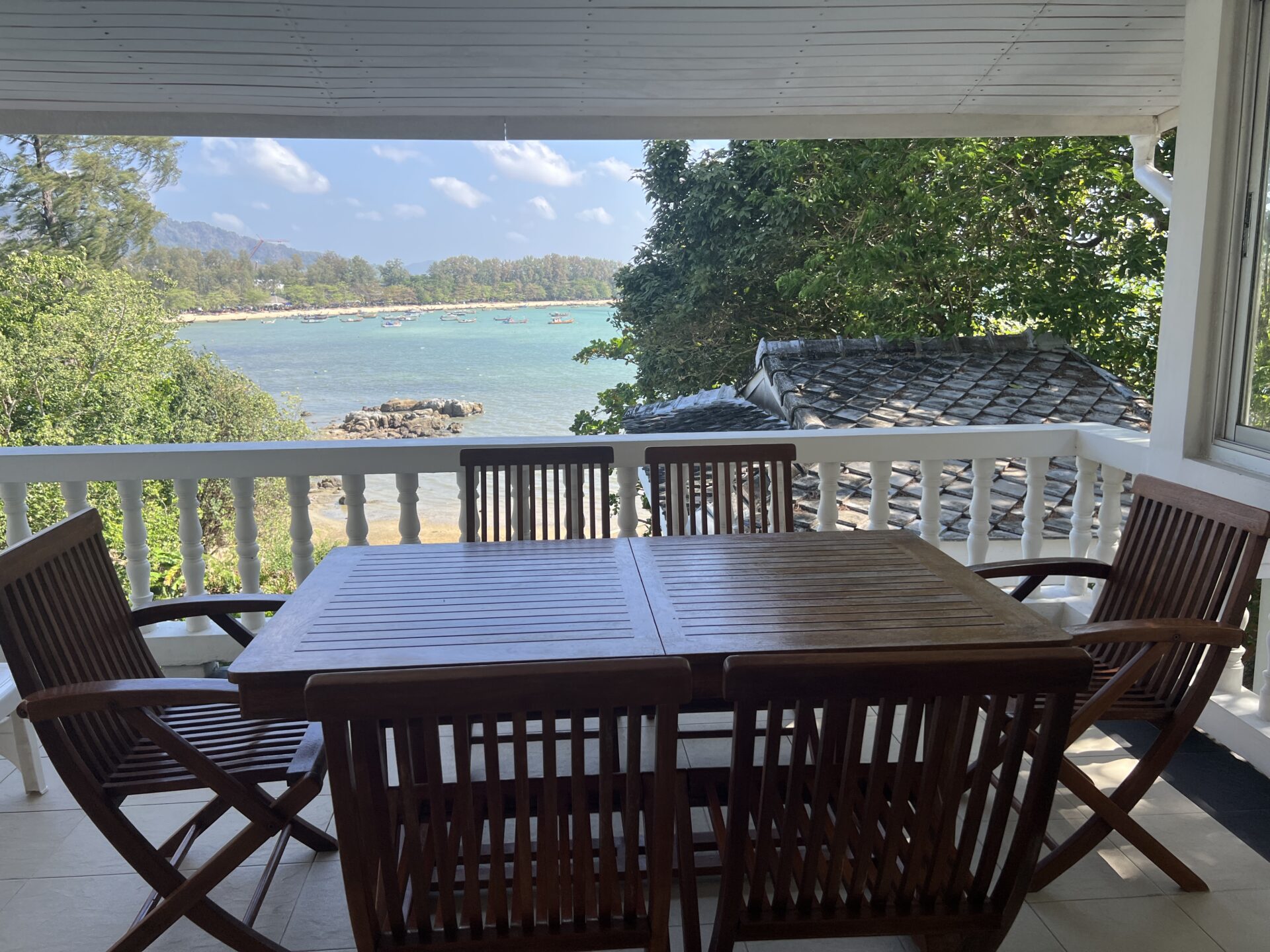 Wooden outdoor dining table and chairs on a covered balustraded patio, overlooking a blue bay with boats and distant beach.