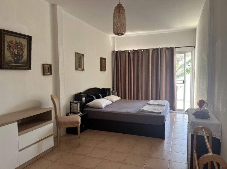 Two single beds pushed together in a bright hotel room with gray bedding, brown curtains, and a wicker pendant light above.
