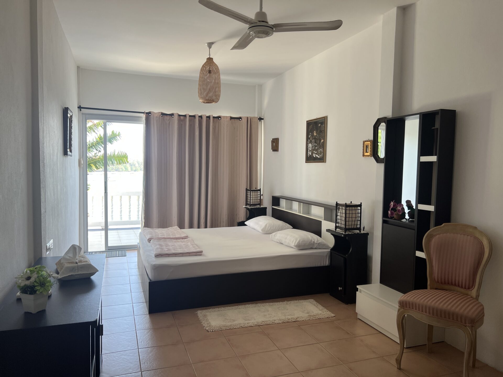 Hotel room with a large dark wood bed, two pillows, and folded linens, near sliding balcony doors with beige curtains
