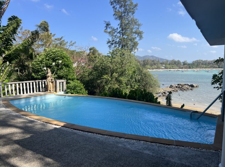 Rectangular blue pool in a tropical garden, with a statue beside a railing, overlooking a rocky shore and a calm bay with boats under a blue sky.