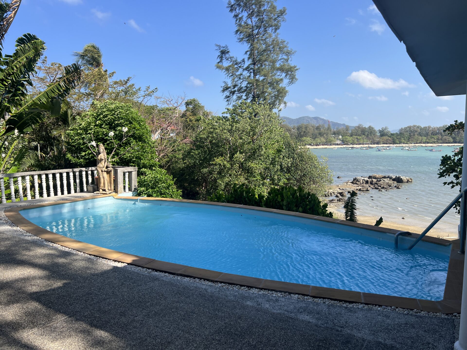 Rectangular blue pool in a tropical garden, with a statue beside a railing, overlooking a rocky shore and a calm bay with boats under a blue sky.