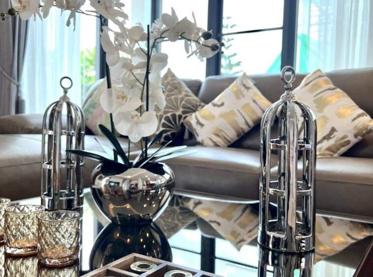 Modern living room centerpiece with white orchid in a shiny silver vase on a glass coffee table, reflective surfaces and patterned cushions in the background.