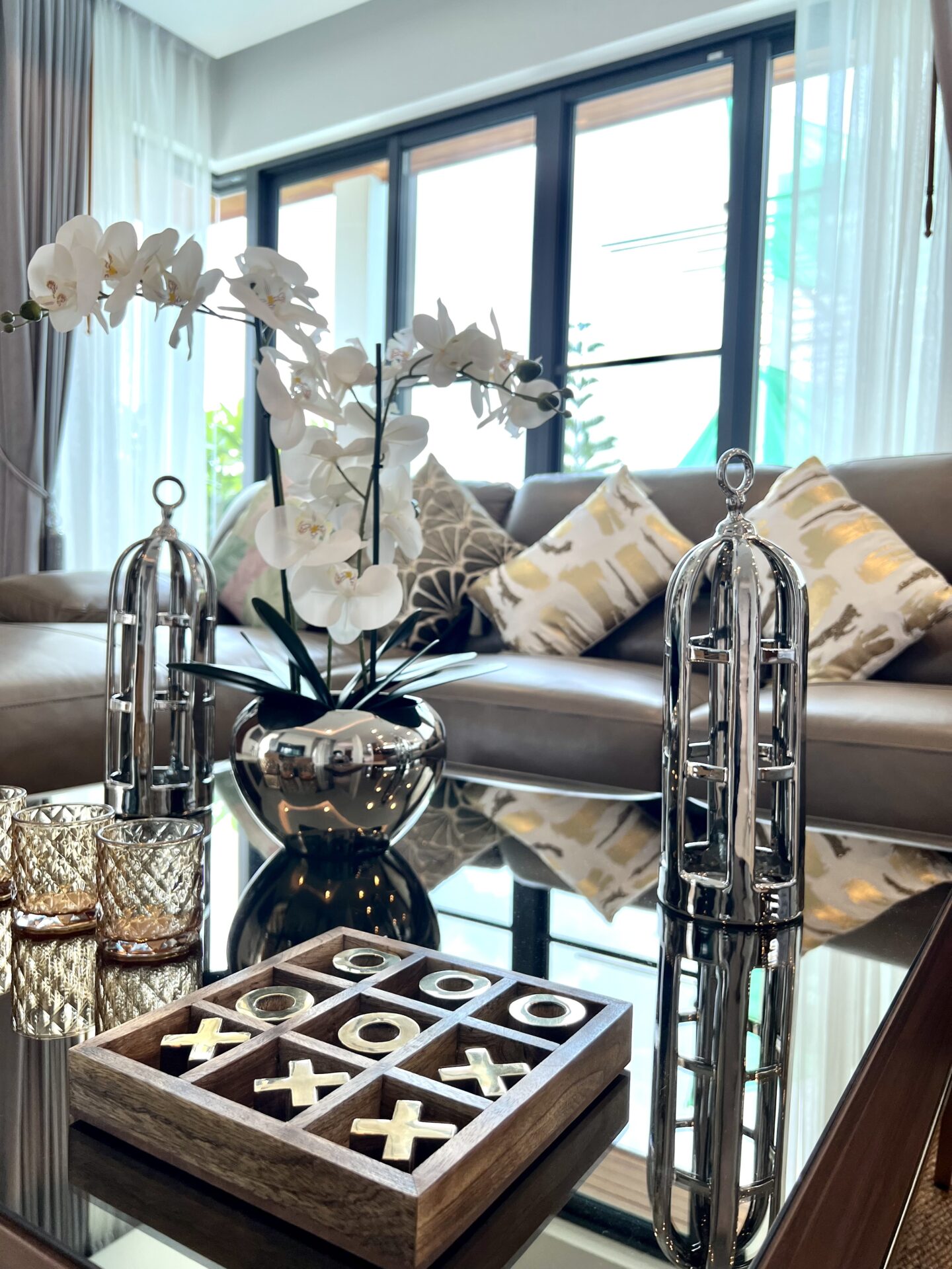 Modern living room centerpiece with white orchid in a shiny silver vase on a glass coffee table, reflective surfaces and patterned cushions in the background.