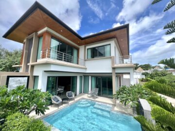 Modern two-story house with wooden overhangs, glass doors, and a rectangular pool in the courtyard, under a bright sky.