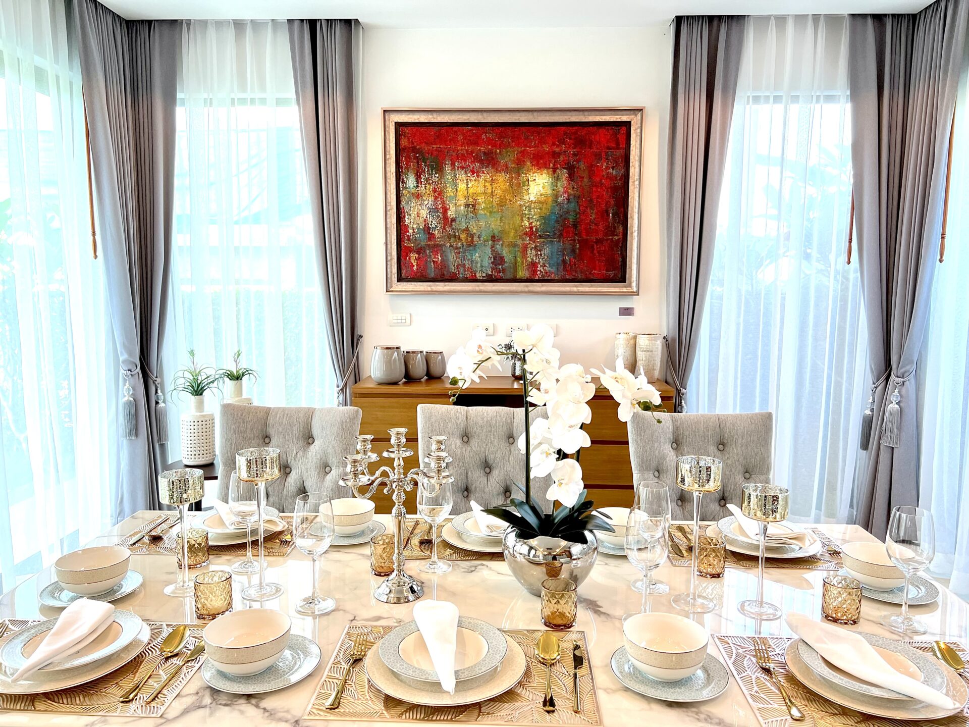 Elegant dining room with a long marble table set for a formal dinner, white napkins, fine china, crystal glassware, and gold-toned candlesticks, framed by gray curtains and a bold abstract painting on the wall.