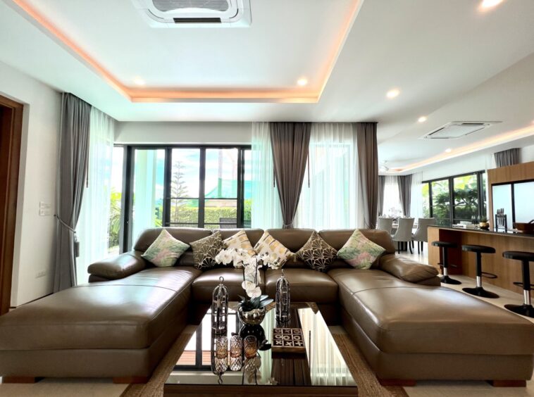 Open-plan living room with a brown L-shaped sectional, decorative cushions, and a glass coffee table near large sliding doors with sheer gray curtains.