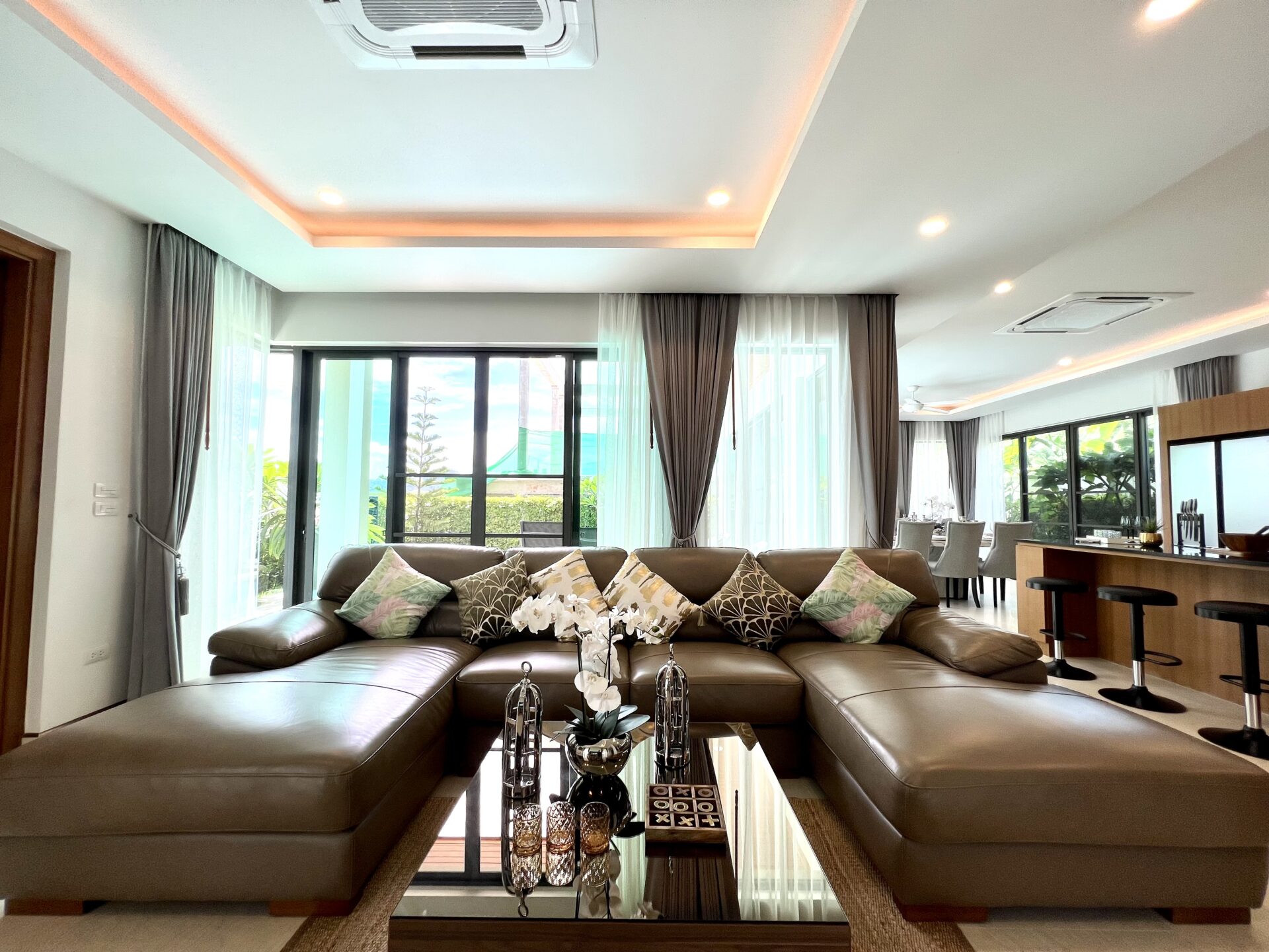 Open-plan living room with a brown L-shaped sectional, decorative cushions, and a glass coffee table near large sliding doors with sheer gray curtains.