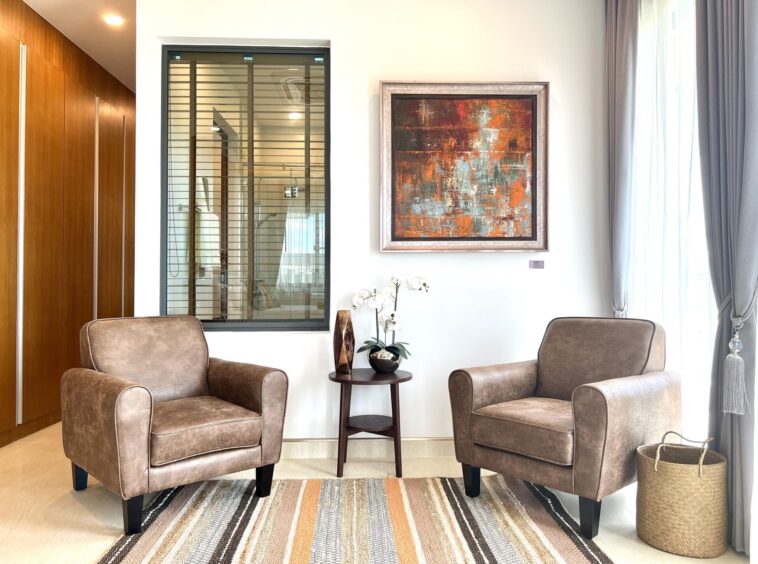 Two beige upholstered armchairs face each other with a round side table and abstract painting on the wall above.