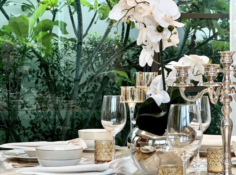 Elegant dining table set with a white orchid centerpiece, crystal glasses, and gold candle holders beside a window with greenery behind it.