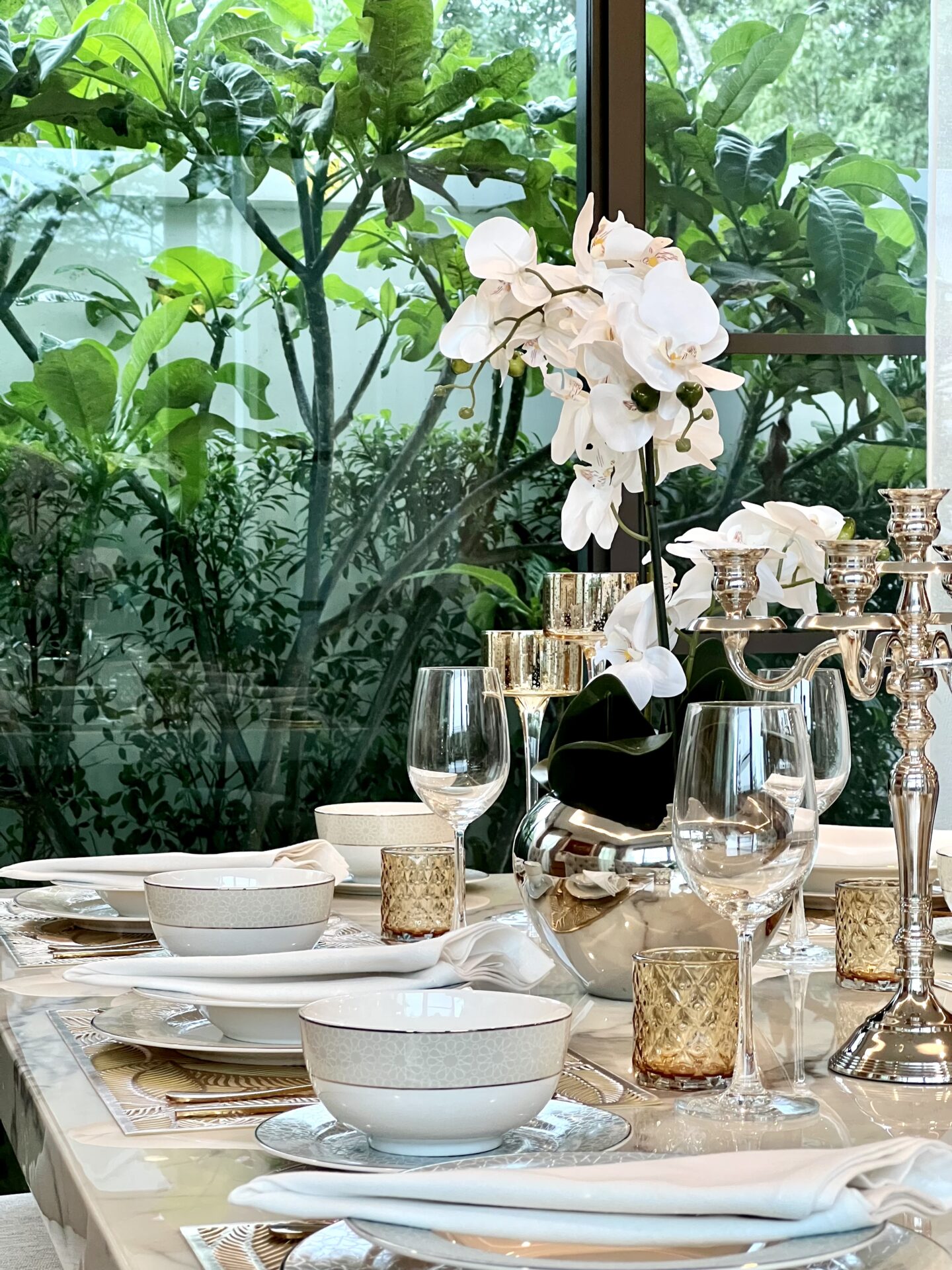 Elegant dining table set with a white orchid centerpiece, crystal glasses, and gold candle holders beside a window with greenery behind it.