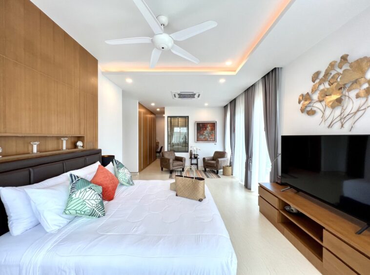 Modern hotel suite: king bed with decorative pillows, wood panel headboard, and a living area with two chairs and a large TV.