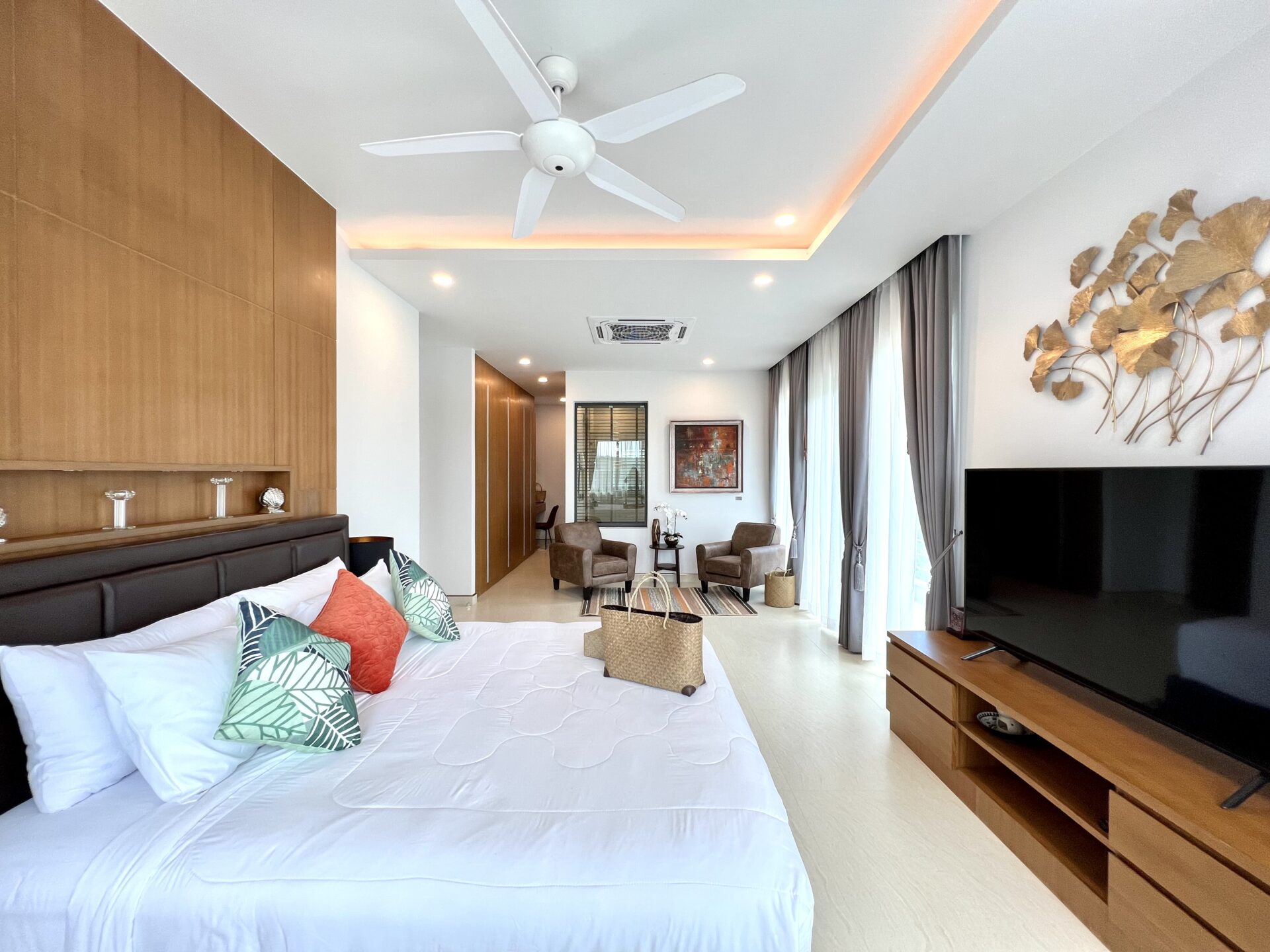 Modern hotel suite: king bed with decorative pillows, wood panel headboard, and a living area with two chairs and a large TV.